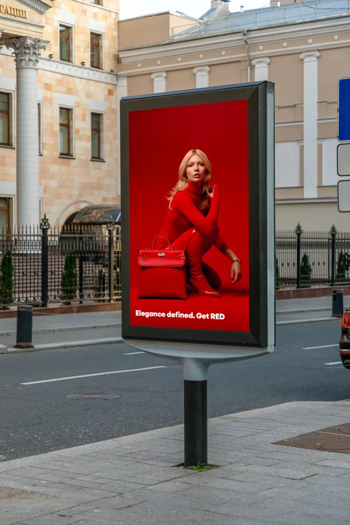 An outdoor advertisement billboard featuring a fashion ad. The poster shows a woman dressed in a stylish red outfit, sitting elegantly, with a red handbag next to her. The background is solid red, making the image and text stand out. The text on the poster reads, "Elegance defined. Get RED." The billboard is located on a city street with buildings and a fence visible in the background.