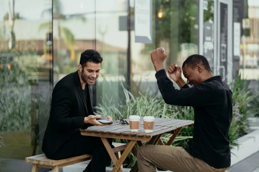 Two men sitting at an outdoor café table, both enjoying coffee. One man, on the left, is holding a smartphone and smiling, while the other man, on the right, is raising his arms in a celebratory gesture. The setting includes a glass window behind them and greenery surrounding the area, creating a pleasant outdoor ambiance.